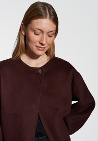 Burgundy wool jacket with a round neckline, front button closure, and side pockets. Smooth texture, loose fit, and minimalistic design.