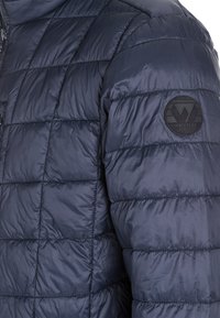 Marinblå puffjacka med quiltat mönster, slät yta och en rund svart logotyp på vänster ärm.