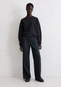 Donkergrijze oversized sweatshirt met reliëflogo, gecombineerd met Hoge taille, wijde donkergroene broek en zwarte schoenen. Soepele texturen.