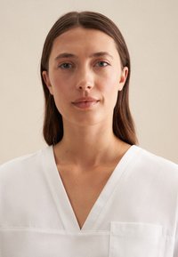 Blusa bianca a V con una vestibilità morbida, caratterizzata da una tasca sul petto e una texture del tessuto liscia. Design semplice con linee pulite.
