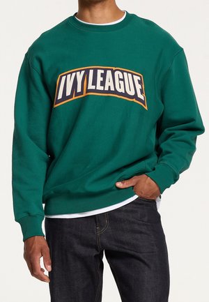 Uomo che indossa una felpa verde con la scritta "IVY LEAGUE" in grandi lettere bianche contornate di blu navy e arancione, abbinata a jeans scuri.