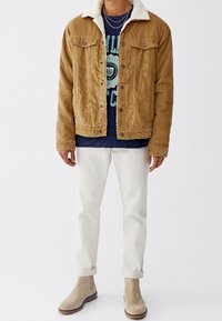 Bruine corduroyjas met een witte nepbontkraag, gelaagd over een marineblauw graphic t-shirt, gecombineerd met witte jeans en tan enkellaarzen.