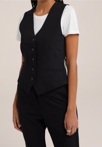 Czarna dopasowana kamizelka o skroju dopasowanym, z pięcioma guzikami z przodu, wyłamiami oraz dwoma przednimi kieszeniami. Nosi się ją na białym t-shircie.