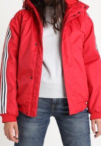 Rød polstret jakke med hætte, med hvide og sorte lodrette striber på ærmerne, trykknaplukninger og en let glans i materialet.