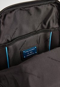 Interno della borsa Piquadro nera con tasca con zip, accenti blu e etichetta con logo. Texture del tessuto liscia con motivi di branding sottili.