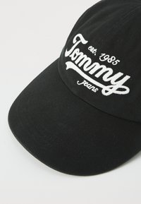 Casquette de baseball noire avec un bord incurvé, ornée du texte brodé en blanc "Tommy Jeans" et de l'année d'établissement, 1985, au-dessus du logo.