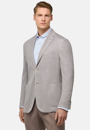 Hellgrauer maßgeschneiderter Blazer mit subtiler Textur, zwei Fronttaschen, Ein-Knopf-Verschluss und Reverskragen. Getragen über einem hellblauen Hemd.