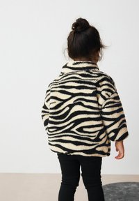 Gestreifte, flauschige Fleecejacke in Beige und Schwarz, mit einem Kragen und weiten Ärmeln, kombiniert mit schwarzen Hosen, von hinten betrachtet.