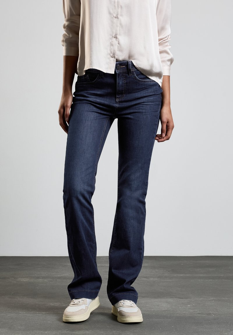 Jean foncé avec une coupe slim, comprenant des poches avant, une taille classique et des coutures discrètes. Associé à des baskets de couleur claire.