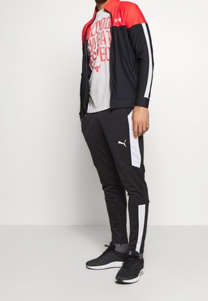 Mężczyzna ubrany w czarno-białe spodnie dresowe Puma, czarno-czerwoną kurtkę Under Armour, szary t-shirt z grafiką oraz czarne tenisówki.