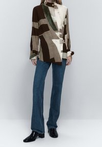 Blazer à motifs marron et crème avec un col haut, associé à un jean évasé en denim bleu et des bottines noires brillantes.