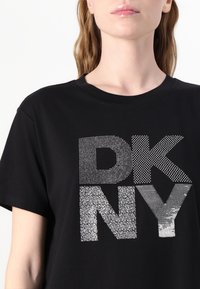 Juodas medvilninis marškinėlis su dideliu "DKNY" logotipu sidabro spalvos strazdanais, demonstruojantis įvairius tekstūras ir grafikos dizainą. Apvalus iškirpimas, trumpomis rankovėmis.