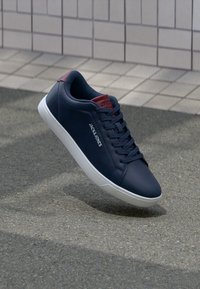 Marineblaue Jack & Jones Sneaker mit weißer Sohle und roten Akzenten, schwebend über strukturiertem Asphalt in der Nähe einer gefliesten Wand.