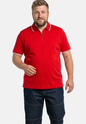 Brodaty mężczyzna w czerwonej polo z krótkim rękawem i ciemnoniebieskich jeansach stoi na tle jednolitego jasnego tła.