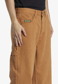 Pantalones cargo de color caramelo hechos de tejido resistente, con un bolsillo de parche y detalles de costura visibles, con etiqueta de la marca en la parte delantera.