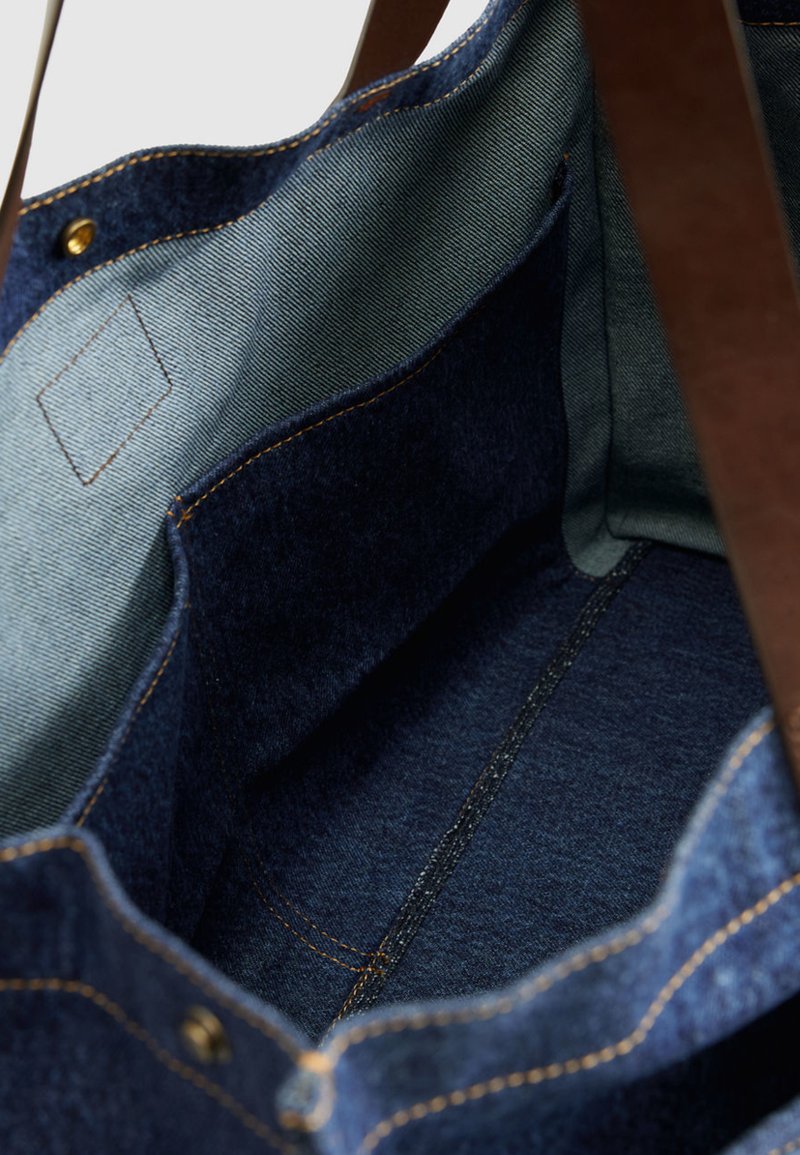 Innenansicht einer Denim-Tragetasche mit großem zentralem Fach, Stofftasche und braunen Leder-Schulterriemen.