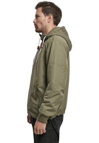 Veste à capuche vert olive avec une poche zippée, des poignets élastiques et des accents à cordon de serrage. Tissu léger avec une texture lisse.