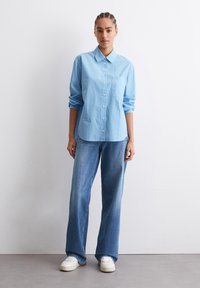 Camicia button-up a righe blu con colletto, abbinata a jeans a gamba larga azzurri e sneakers bianche. Tessuto liscio, silhouette rilassata.