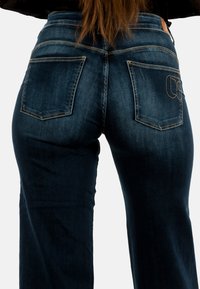 Donkerblauwe denim jeans met een hoge taille, uitgerust met twee achterzakken en contrasterende stiksels. De stof lijkt soepel met een lichte glans.