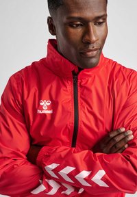 Mann in roter Hummel-Jacke mit weißen Chevron-Streifen auf den Ärmeln, steht mit verschränkten Armen und schaut nachdenklich nach unten.