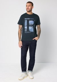 Svart bomulls-t-shirt med texten "ROUGH PEAK". Har korta ärmar och rund halsringning. Matchas med mörkblå jeans och vita sneakers.
