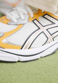 Fehér sportcipő narancssárga és fekete részletekkel, hálós és szintetikus anyagokból készült, ívelt formájú és texturált felületekkel. Talpa fekete gumitalpból készült.