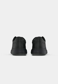 Par de zapatillas negras de Calvin Klein mostradas desde atrás, con el logo elevado de la marca en los talones y un discreto patrón de monograma en los laterales.