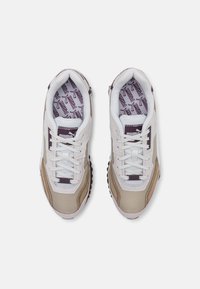 Sneaker con una combinazione di materiali bianchi, beige e marrone scuro. Sono visibili dettagli testurizzati, design con lacci e suole interne a motivi.