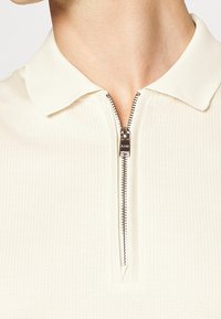 Krémová polo košile s texturovanou tkaninou, vybavená předním zipovým zapínáním a klasickým límcem. Detail stříbrného zipového kování.