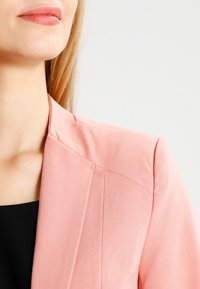 Blazer rose avec épaules structurées, tissu lisse et détails de couture à l'avant. Porté sur un haut noir, mettant en valeur un décolleté net.