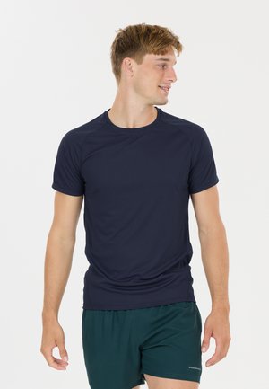 Ung mand iført navyblå kortærmet sportstrøje og mørkegrønne shorts, kigger mod højre mod en ensartet lys baggrund.