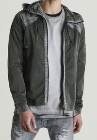 Lekka oliwkowo-zielona kurtka z kapturem i kontrastowymi szarymi akcentami. Posiada przedni zamek błyskawiczny, dwie boczne kieszenie i jest częściowo rozpięta.