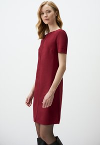 Burgunderfarbenes Kurzarmkleid mit Rundhalsausschnitt, geradem Schnitt, seitlichen Taschen und glatter Textur, ergänzt durch schwarze knöchelhigh Stiefel.