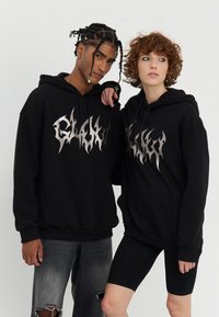 Zwarte hoodies hebben een zilveren vlamstijl logo op de borst. De stof voelt zacht aan, heeft een ontspannen pasvorm en voorzakken.