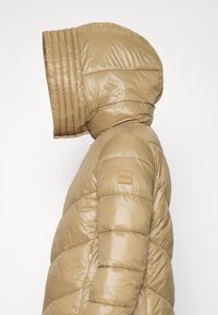 Giacca puffer beige con texture trapuntata, grande cappuccio strutturato e un piccolo patch logo sulla spalla. Presenta una chiusura con zip.