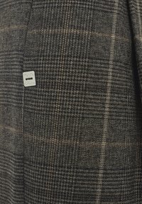 Tissu en mélange de laine dans un motif à carreaux gris foncé et marron avec une petite étiquette en métal. Surface texturée avec une densité de fil variable.