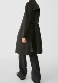 Czarny trench z paskiem w talii, podwójnymi guzikami i detalem na plecach, w połączeniu z szerokimi ciemnymi dżinsami.