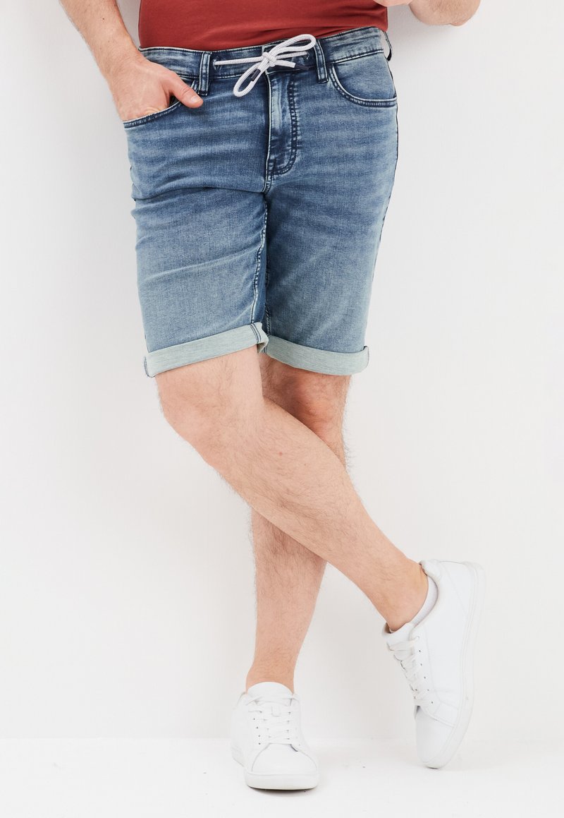 Shorts de mezclilla en azul claro con dobladillos enrollados, que cuentan con una cintura ajustable con cordón y bolsillos delanteros, combinados con zapatillas blancas.
