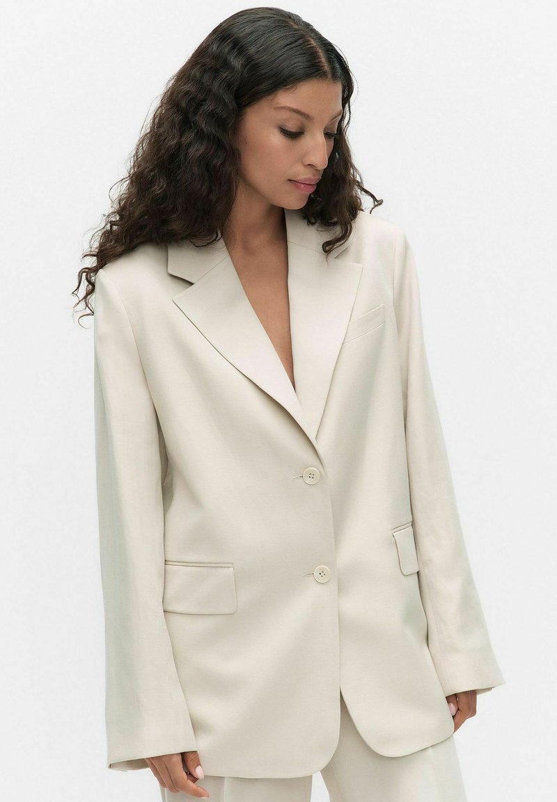 Pitkäkiharainen nainen yllään ylimittainen vaaleanbeige bleiseri, jossa on kaksi nappia ja läppätaskut, katsoo alas vasemmalle.