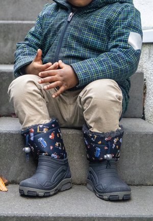 Kind trägt karierten Mantel, beigefarbene Hose und gemusterte Winterstiefel und sitzt auf einer Außentreppe aus Beton mit verschränkten Händen.