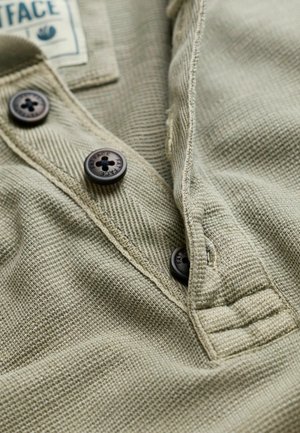 Khaki texturált szövet gombos patenttal, három sötét gombraval és egy kis címkével. Varrt szélek és enyhe foszlás részlet.