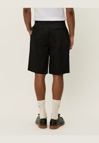 Pantaloni shorts neri con una texture liscia, taglio a lunghezza ginocchio, due tasche posteriori, abbinati a calze crema e scarpe stringate nere.