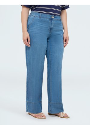 Vrouw draagt lichtblauwe wijde jeans en beige sandalen, staande met ontspannen handen langs haar zij.