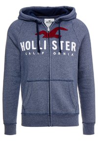 Marinblå zip-hoodie i mjukt tyg med en stor röd broderad logga, två framfickor och en dragsko på huvan.