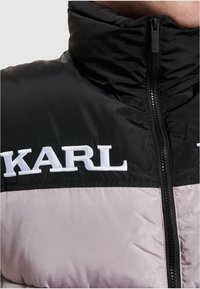 Zateplená bunda s vysokým golierom, čierna horná časť a svetlo šedá dolná časť. Na prsiach vyšívané "KARL" v bielej farbe.