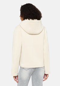 Cremefarbene Kapuzenjacke mit strukturiertem Design, glatter Textur und geradem Schnitt, kombiniert mit hellgrauen, eng anliegenden Jeans.