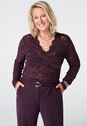 Blonde vrouw die glimlacht, draagt een donkerpaarse kanten top met lange mouwen, bijpassende broek met riem en gelaagde gouden kettingen, handen in de zakken.