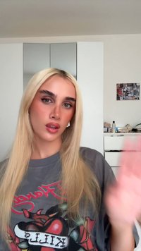 Blont hår, långt och rakt. Grå t-shirt med djärva röda grafik och en färgglad design. Neutral bakgrund med vit köksinredning.