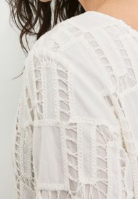 Blusa blanca con intrincados patrones tejidos, detalles de punto abierto y acentos texturizados, que presenta un ajuste relajado y un cuello redondo.