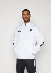 Hvid zip-up hoodie lavet af blødt stof, med sorte Adidas-striber på ærmerne og Juventus-logoet på venstre bryst. Simpelt, moderne design.